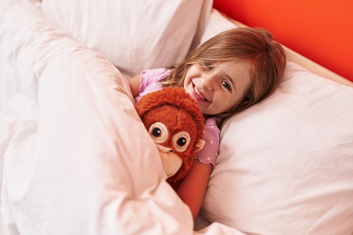 Adorable hispanic girl hugging monkey doll lying on bed at bedroom