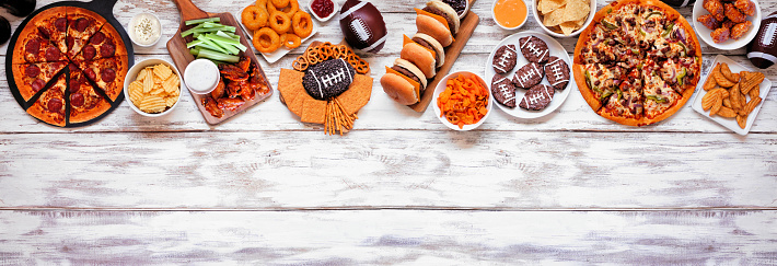 Super Bowl or football theme food top border. Pizza, hamburgers, wings, snacks and sides. Above view on a white wood banner background.