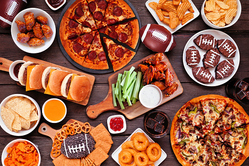 Super Bowl or football theme food table scene. Pizza, hamburgers, wings, snacks and sides. Overhead view on a dark wood background.