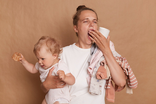 Portrait of sleepy overworked woman wearing white T- shirt holding her baby daughter in hands isolated over brown background, holding coffee cup, yawning, covering mouth with palm.