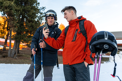 Two young boys standing in ski clothes looking at the cell phone of one of the boys. They are talking and smiling. In the background you can see the snow slopes
