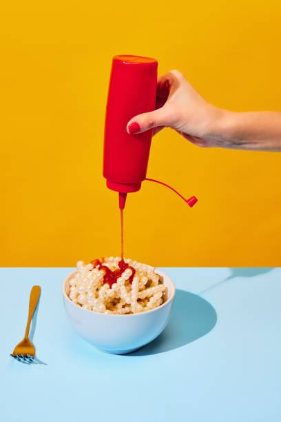 main féminine versant du ketchup dans un bol avec des colliers symbolisant les pâtes, des nouilles sur une nappe bleue sur fond jaune vif. photographie pop art culinaire. - eating senior adult color image spaghetti photos et images de collection