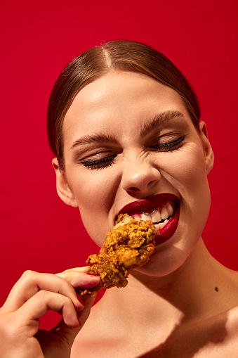 Young woman with red lipstick eating fried chicken, nuggets over vivid red background. Fast food lover. Food pop art photography. Complementary colors. Junk food. Copy space for ad, text