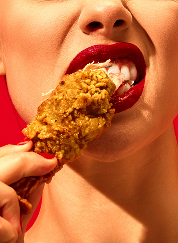 Close-up cropped image of young woman eating fried chicken, nuggets over vivid red background. Spicy taste. Food pop art photography. Complementary colors. Copy space for ad, text