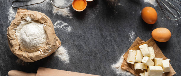 istock Ingredients for cooking baking dough, paper bag with flour, butter and eggs, rolling pin and sieve on dark background top view 1455072461
