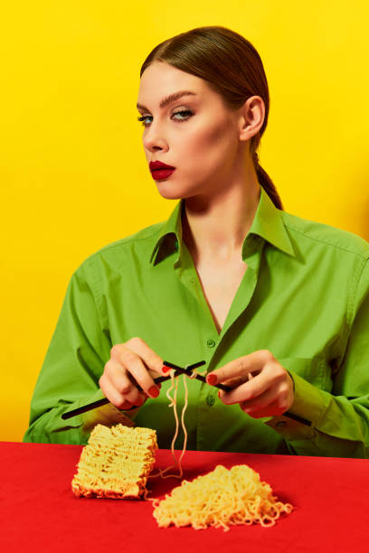 woman with emotionless face knitting instant noodles on blue table on vivid red tablecloth over yellow background. food pop art photography. - eating senior adult color image spaghetti imagens e fotografias de stock