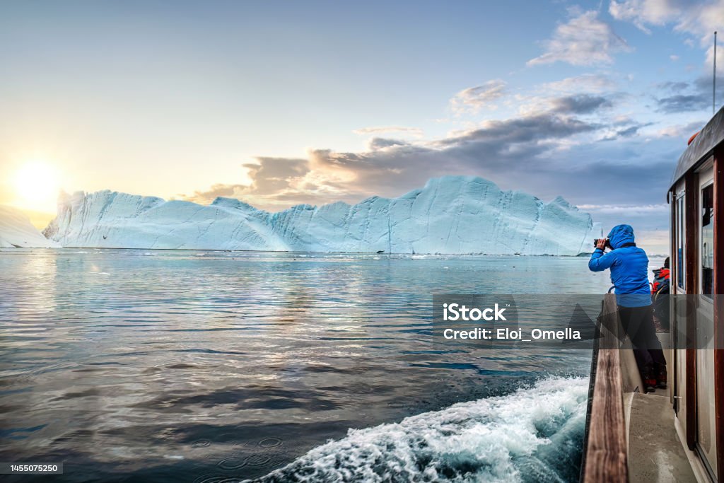 Travel in arctic landscape nature with icebergs,  tourist man explorer from a boat at sunset. Greenland Tourists in a boat in front of an iceberg taking photos at midnight sun sunset, Ilulissat Icefjord, Disko bay, Greenland Ilulissat Stock Photo