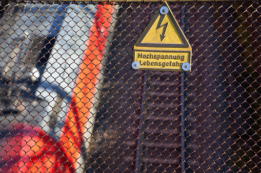 Sign on a old rusty fence saying: High voltage, danger to life ( german: Hochspannung Lebensgefahr ). Rails in the background, red train passes by. Closeup, top view.