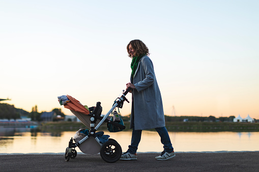 Young adult mother walking at sunset with baby carriage in park
