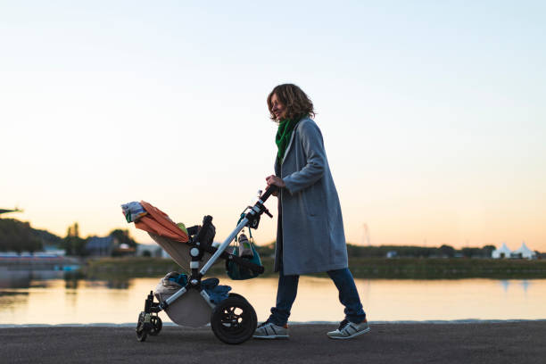Young adult mother walking at sunset with baby carriage in park stock photo