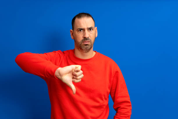 istock Bearded hispanic man wearing red sweater sulking shows thumb down with negative attitude, isolated over blue background. 1455082071