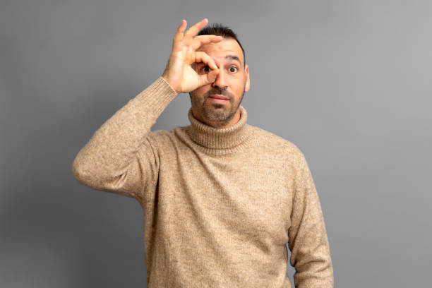 istock Kind guy making ok gesture looking through fingers. Bearded latino man wearing a turtleneck sweater making the ok gesture over his eye isolated over gray background. 1455082074