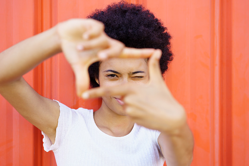 Glad African American female in casual wear winking and looking at camera through frame gesture on red background in city