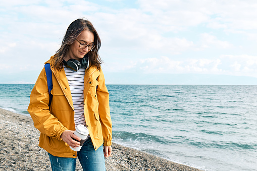 Smiling woman in yellow jacket with backpack and coffee mug portable walking along sea coast and enjoying the sunny day. Concept of hiking and activity in the low season.