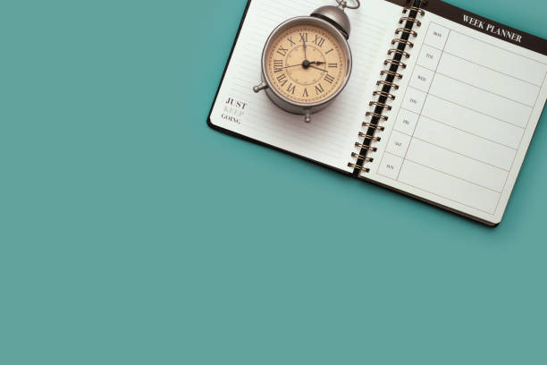 Overhead flat lay of a calendar planner notebook and a vintage alarm clock against a teal background.
