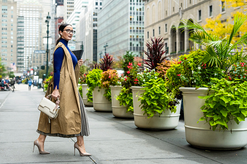 Beautiful woman in a beige sleeveless coat, pleated skirt, turtleneck sweater, high heels and with a handbag seen exiting the building in Manhattan, New York, while using her wireless headphones and holding a laptop after finishing work.