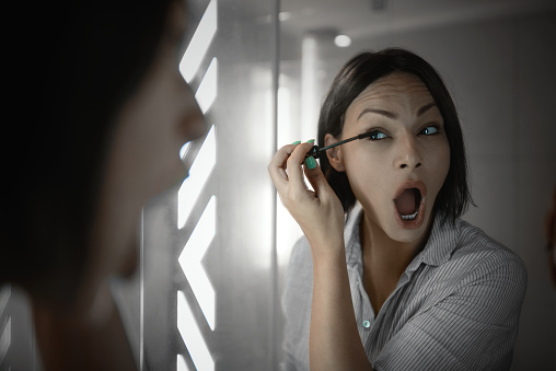 Beautiful woman applying mascara in front of the mirror in the bathroom. She's getting ready for work.