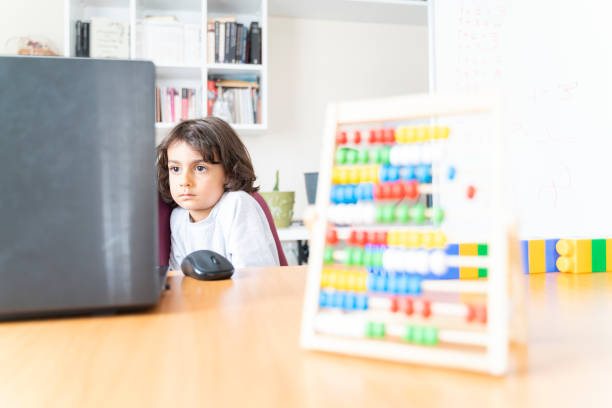 little boy spending time in the study of the house. There is a bookshelf and a whiteboard in the back. kid watching video on laptop. Shot with a full-frame camera in daylight.