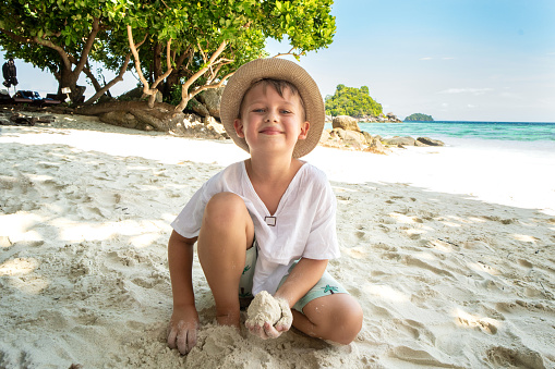 Little happy boy in summer hat playing with sand on the tropical beach, looking at the camnera with big smile on his face. Beautiful landscape. Blue sky and sea water background. Kid on vacation.