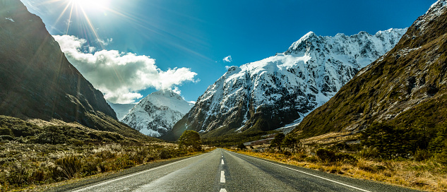 A road leading towards rugged snowcapped mountains with blue sky and clouds. Sun rays in top corner of image.