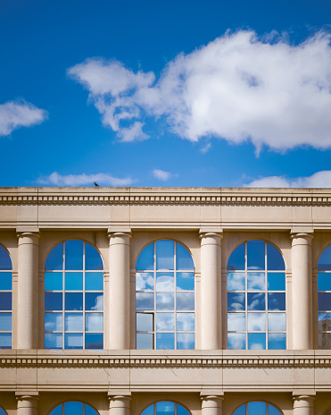 On the photo, there is a building with a Greek-style architecture, featuring columns, a triangular pediment, and a symmetrical facade. The building has windows that reflect the clouds in the sky