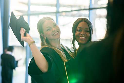 Two University graduates pose together for a portrait as a classmate takes a photo on her cell phone.  They are each dressed in formal black gowns and graduation caps as they smile with pride and excitement.