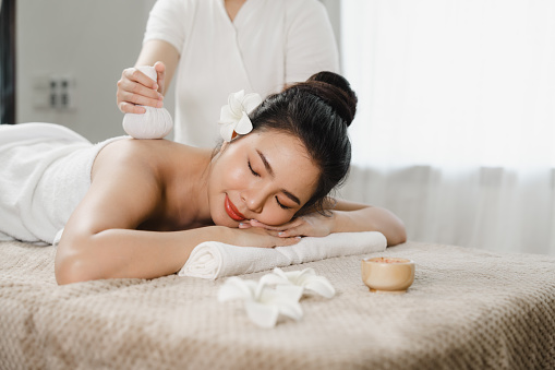 Relaxed pretty asian young woman enjoying remedial body massage done by professional masseur in spa room, with herbal compress balls in therapy spa salon.
