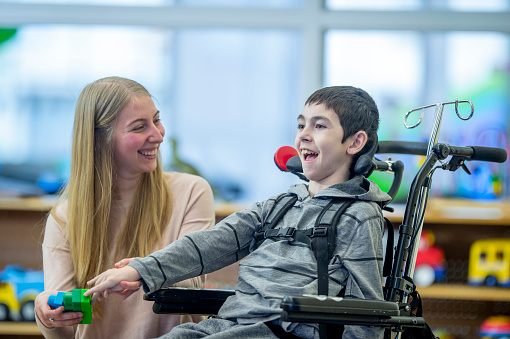 An Elementary student with Multiple Sclerosis sits in his wheelchairs as his teachers helps him with some gross motor skills.  He is dressed casually and they are using colorful blocks to work on strengthening his co-ordination.