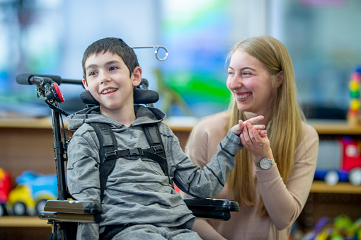 An Elementary student with Multiple Sclerosis sits in his wheelchairs as his teachers helps him with some gross motor skills.  He is dressed casually and they are using colorful blocks to work on strengthening his co-ordination.