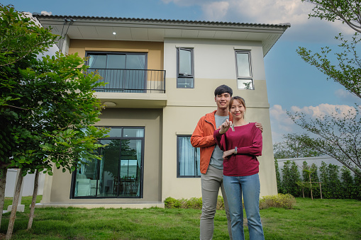 Portrait of young Asian couple received house keys and standing outside at new house, Loan and mortgage, Homeowners and New home concept, buy or sale and moving home, first property purchase and Life event, looking at camera