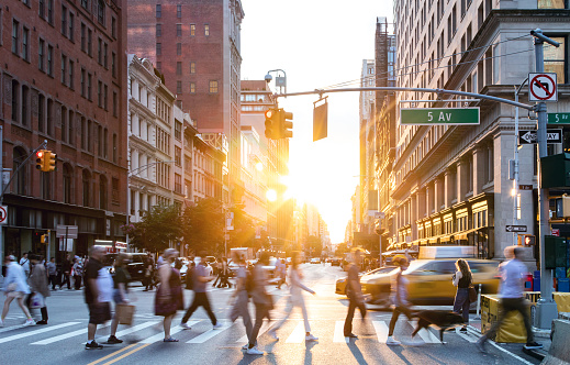 People and cars in a busy intersection on 5th Avenue and 23rd Street in New York City with sunlight shining between background buildings