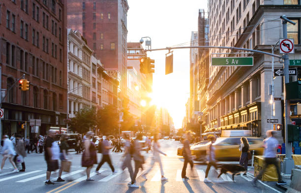 People and cars in a busy intersection on 5th Avenue and 23rd Street in New York City with sunlight shining between background buildings stock photo