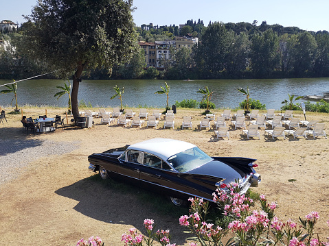 Sun chairs in summer on the Arno riverbank in Florence, Tuscany. An old american car is parked on.