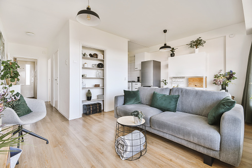 a living room with wood flooring and grey couch in the center of the room is an open kitchen area