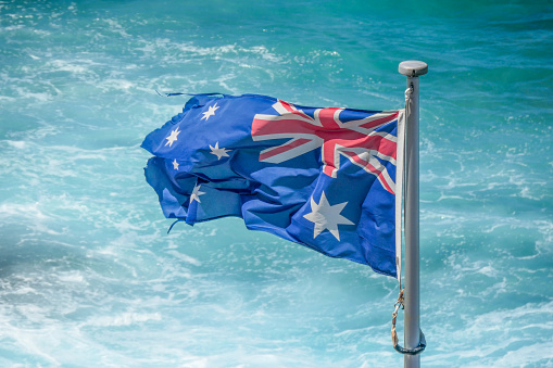 A stained, frayed and torn Australian flag flies at Bondi Beach surrounded by turbulent waters of the Pacific Ocean.  This image was taken on a sunny and windy afternoon in summer.