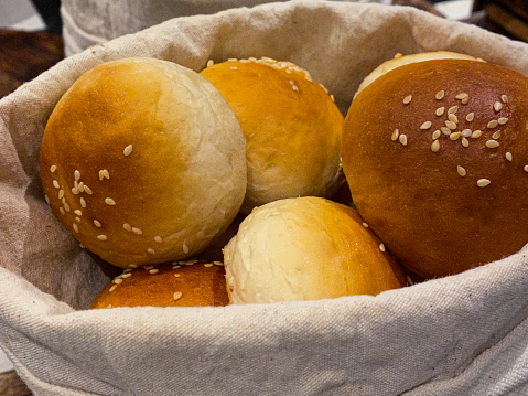 Stock photo showing close-up, elevated view of bakery shelf with linen lined basket of artisan bread rolls topped with sesame seeds. These buns are freshly baked and are ready to be sold.