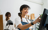 Happy young Asian woman cashier wears an apron and using pos terminal to input orders on coffee shop counter.