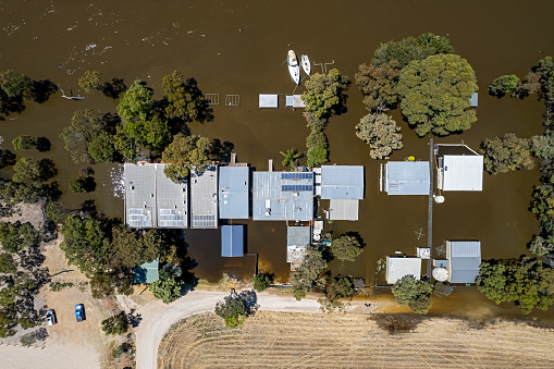 Aerial view looking directly down on a row of treelined flooded houses and holiday shacks surrounded by murky brown floodwaters on River Murray, South Australia; two recreational boats are moored in front of one of the houses. Solar panels, clothes line, sheds, flooded yards.  Town planning issues with residences & vacation rentals built on floodplain. Murray Bridge, Jan 6, 2023
