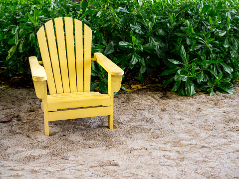 An empty yellow wooden sun chair on the sand beach on green leaves background with copy space. Wood adirondack chair light yellow color with nobody in summer vacation.