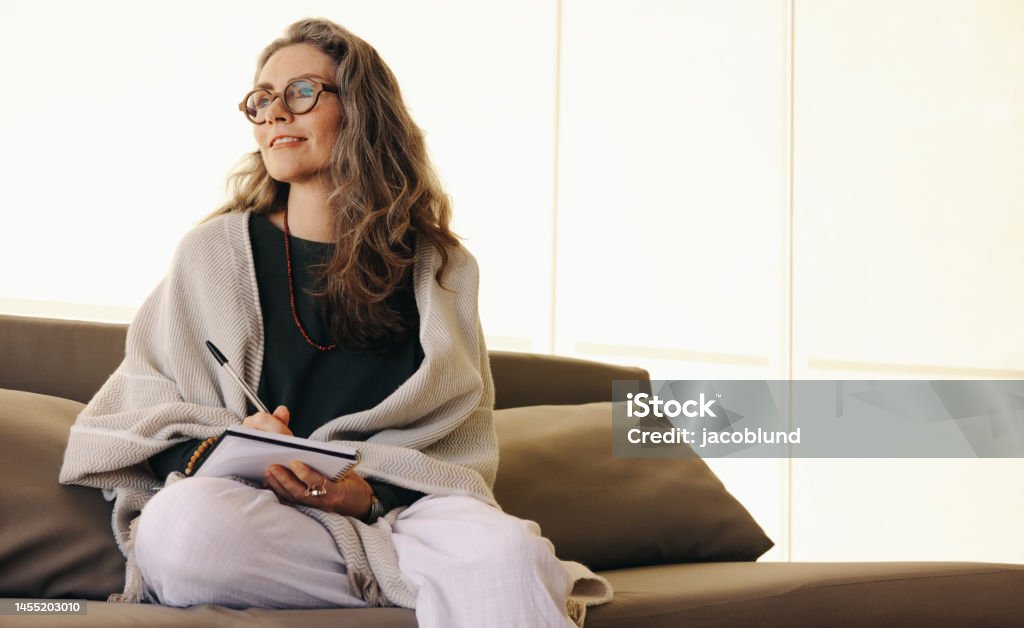 Journaling and self-awareness Journaling and self-awareness. Mature woman looking away thoughtfully while writing in her journal at home. Senior woman writing down her thoughts while sitting on a couch. Note Pad Stock Photo