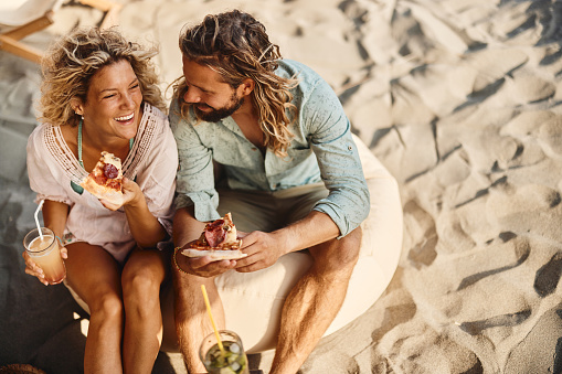 High angle view of happy couple communicating while eating pizza in summer day on the beach. Copy space.