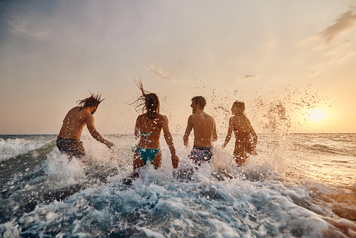 Rear view of group of playful friends having fun while rushing into sea at sunset. Copy space.