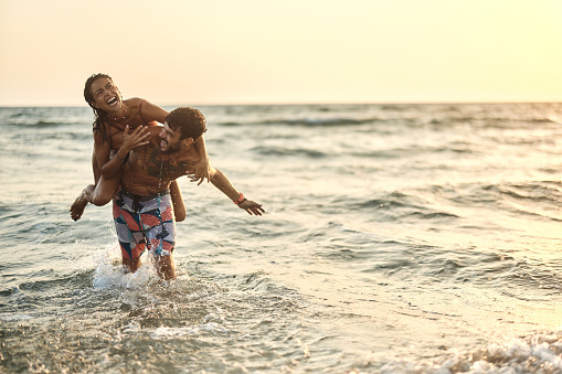 Young playful couple having fun while piggybacking in summer sunset at sea. Copy space.