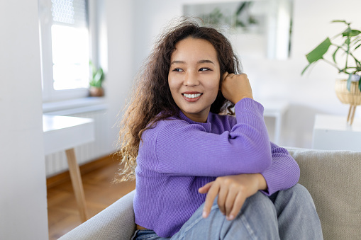 Happy Asian woman sitting on sofa at home and looking at camera. Portrait of comfortable woman in winter clothes relaxing on sofa . Portrait of beautiful woman smiling and relaxing during autumn.