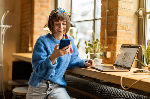 Young cheerful woman works on phone and laptop while sitting by the window with a coffee drink at modern cafe. Concept of remote work from public place, digital freelance and modern lifestyle