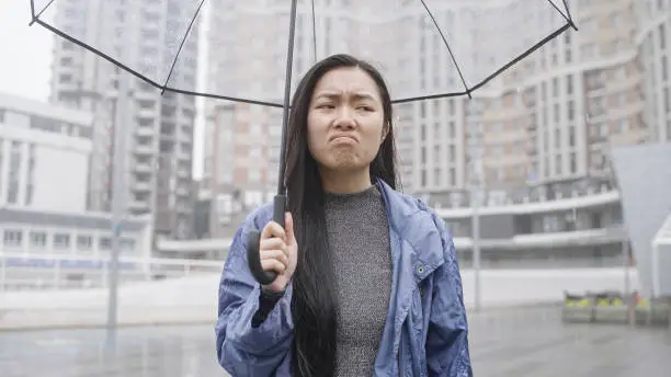 Upset asian woman standing in rain under umbrella, ruined plans, bad mood, melancholy