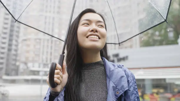 Happy asian woman smiling, enjoying rain, walking city with transparent umbrella