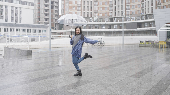 Positive cheerful asian woman dancing outside in rain, hiding under big umbrella