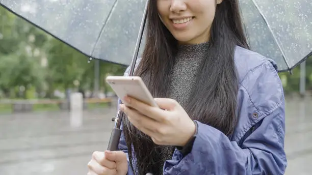 Young asian woman smiling, chatting with friend on smartphone, standing in rain