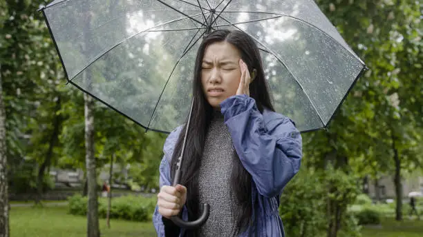 Frustrated asian woman feeling headache standing in rain, weather-related pain